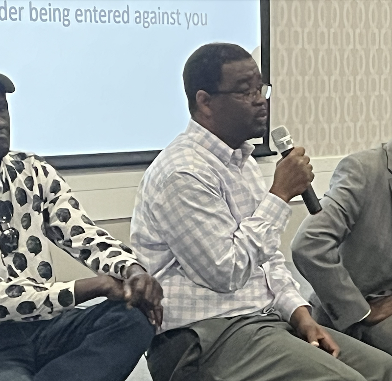 Three Black men sit on a stage and address a community meeting, one holds a mic